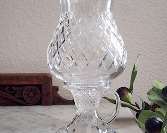 May include: A clear, cut-glass candle holder with a diamond pattern on the bulbous body. It features a handle and sits on a decorative base. The candle holder is designed to hold a single candle.