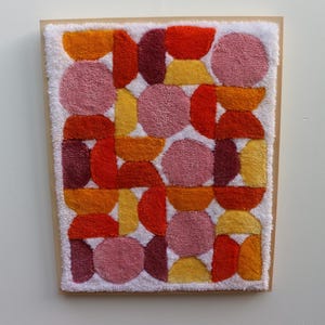 Peut inclure: Une œuvre d'art textile encadrée avec un motif géométrique. L'œuvre présente un fond blanc avec un motif de formes orange, rouge, jaune et rose. Les formes comprennent des cercles et des demi-cercles. Le cadre est de couleur marron clair.