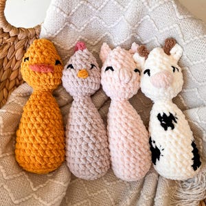 May include: Four handmade crochet farm animal toys: a yellow duck, a lavender chicken, a pink pig, and a black and white cow. The toys are placed in a woven basket on a textured blanket. Each toy has embroidered facial features.