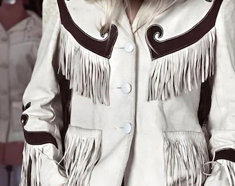 Op de afbeelding: Crème leren jas met bruine accenten en franjes. De jas heeft een knoopsluiting, decoratieve schouder- en mouwaccenten en franjes op de borst, zakken en mouwen. Een cowboyhoed is gedeeltelijk zichtbaar.