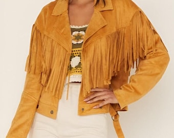May include: Mustard-colored suede jacket with fringe detailing across the chest, back, and sleeves. The jacket features a notched collar, button closures, and a belt detail at the waist. The jacket is paired with a patterned top and white pants.