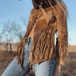 May include: Brown suede jacket with star accents and fringe detailing. The jacket features a lace-up front and is paired with blue jeans. The sleeves have fringe and beaded accents. A western-inspired fashion piece.