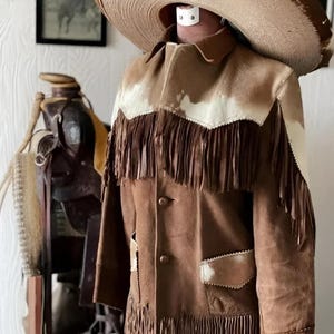 Puede incluir: Chaqueta de ante marrón con flecos, detalles de piel de vaca blanca y marrón y dos bolsillos delanteros. La chaqueta se exhibe con un gran sombrero color canela. Una silla de montar es visible en el fondo.