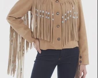 May include: Tan suede jacket with long fringe and turquoise accents. The jacket has a button-down front and long sleeves. The fringe is along the front and sleeves. The model is wearing dark blue jeans.