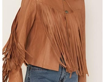 May include: A tan leather jacket with long fringe detailing on the sleeves and front. The jacket has a round collar and two button closures. The model is wearing blue jeans.