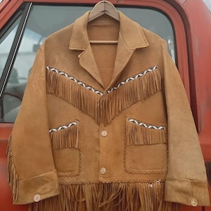 Könnte beinhalten: Eine hellbraune Wildlederjacke mit Fransen. Die Jacke hat einen Reverskragen, Knopfverschlüsse und Taschen. Die Fransen befinden sich an der Vorderseite, den Taschen und den Ärmeln. Die Jacke hängt an einem Holzbügel.