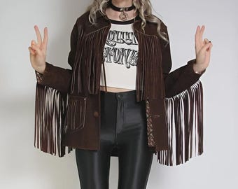 May include: A brown suede jacket with long fringe detailing on the sleeves and chest. The jacket has a button closure and pockets. A white t-shirt with the text "STONE" is visible underneath. The model is wearing black leather pants.