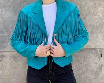 May include: A turquoise suede jacket with long fringe detailing on the sleeves and chest. The jacket has a classic collar, two gold buttons, and a cropped fit. The model is wearing a white shirt and black jeans.