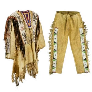 Könnte beinhalten: Ein beigefarbenes Outfit der amerikanischen Ureinwohner, bestehend aus einem Poncho-Oberteil und einer Hose. Das Oberteil weist aufwendige Perlenarbeiten in Weiß, Blau und Rot mit Fransen auf. Die Hose hat Perlenakzente und Fransen an den Seiten, die traditionelle Handwerkskunst zeigen.