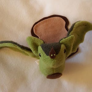 May include: A green plush bat with brown felt wings and a brown felt nose. The bat is sitting on a brown felt background.