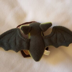May include: A plush green and brown bat toy with large wings. The bat has a stitched face and a small tag on the wing.