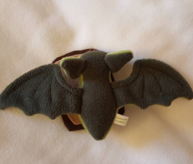 May include: A plush green and brown bat toy with large wings. The bat has a stitched face and a small tag on the wing.