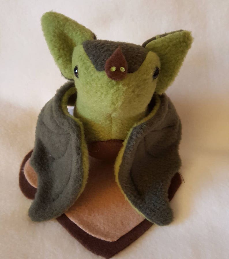 May include: Green and brown plush bat with large ears and wings. The bat is sitting on a brown and tan fabric base.
