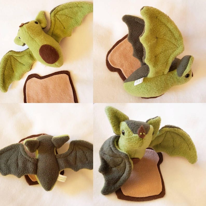 May include: A green and brown plush bat toy with wings spread out. The bat is sitting on a brown piece of fabric that looks like a slice of toast.