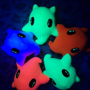 Può includere: Cinque peluche a forma di stella, ognuno con grandi occhi ovali neri e una bocca piccola. I giocattoli sono in tonalità di verde, arancione, rosso e blu. Lo sfondo è blu scuro con un motivo floreale.