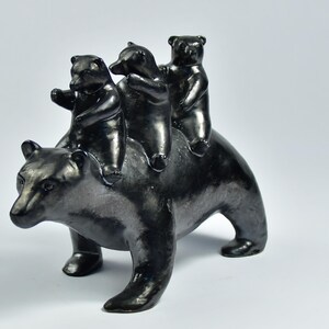 Op de afbeelding: Een zwarte keramische sculptuur van een beer met drie kleinere beren op zijn rug. De beren bevinden zich allemaal in een speelse pose, waarbij de kleinere beren van de rit lijken te genieten. De sculptuur heeft een glanzende afwerking en staat tegen een witte achtergrond.
