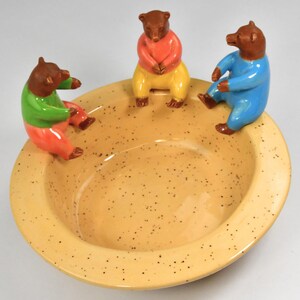 May include: A yellow ceramic bowl with a speckled glaze features three brown bear figurines sitting around the rim. The bears are wearing colorful clothing: green and pink, red and yellow, and blue.