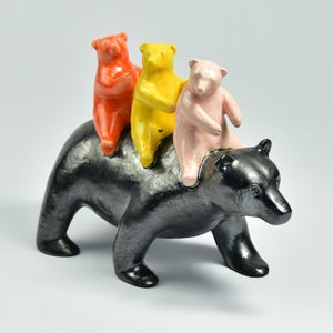 Peut inclure: Sculpture en céramique représentant un ours noir avec trois ours plus petits sur son dos. Les petits ours sont orange, jaune et rose. La sculpture est sur fond blanc.