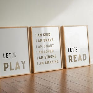 May include: Three framed art prints with positive messages. The first print says "Let's Play" in black and taupe. The second print lists affirmations. The third print says "Let's Read" in black and taupe. The frames are light wood.
