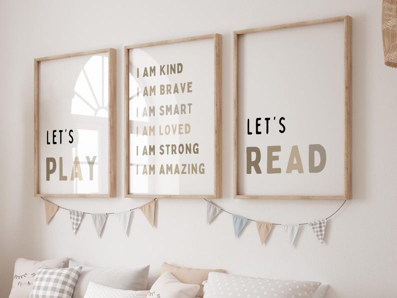 May include: Three framed wall art prints with wooden frames. The prints feature the phrases "LET'S PLAY", "I AM KIND, I AM BRAVE, I AM SMART, I AM LOVED, I AM STRONG, I AM AMAZING", and "LET'S READ" in black and beige lettering. A garland of small flags hangs below.