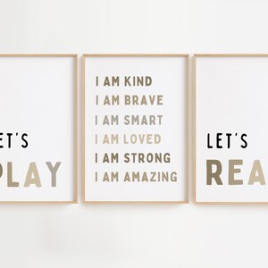 May include: Three framed prints with positive messages. The left print says "LET'S PLAY" in black and taupe. The centre print lists affirmations. The right print says "LET'S READ" in black and taupe. All are in light wood frames.