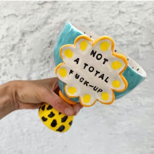Peut inclure: Un bol en céramique avec un intérieur turquoise et un accent en forme de fleur blanc et jaune. L'accent indique "NOT A TOTAL FUCK-UP" en texte noir. La base est jaune avec des taches noires.