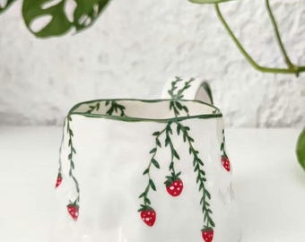 Puede incluir: Taza de cerámica blanca con forma irregular. Decorada con vides verdes y fresas rojas. El borde y el asa presentan diseños de hojas verdes. Un diseño encantador y caprichoso para la cocina o la decoración del hogar.