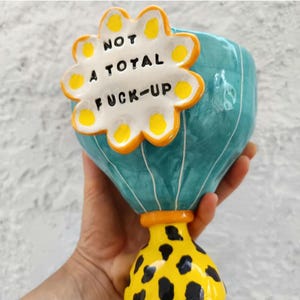 Può includere: Un vaso di ceramica con corpo turchese e base gialla a pois neri. Un accento bianco a forma di fiore con la scritta "NOT A TOTAL FUCK-UP" è attaccato alla parte anteriore del vaso. Il vaso è tenuto da una persona.