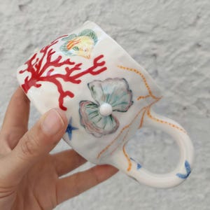 May include: White ceramic mug with a marine theme. The mug features hand-painted coral, a fish, a seashell with a pearl, and starfish. The handle and rim are decorated with blue and orange accents. The mug is held by a person's hand.