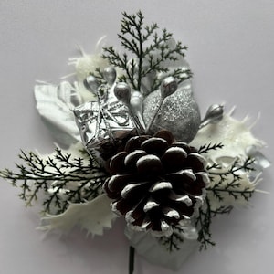 May include: A Christmas floral pick with a brown pinecone, silver baubles, and white leaves. The pick includes sprigs of green and silver foliage, and a silver gift box. The stem is green.
