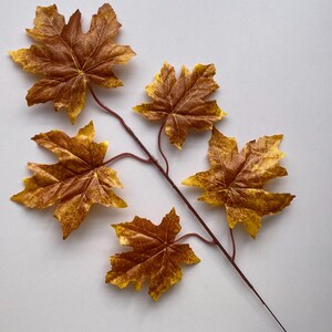 May include: A branch of artificial autumn leaves with five brown maple leaves. The leaves are arranged in a fan shape and have a realistic texture.