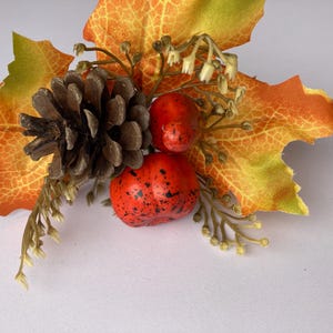 May include: A decorative autumn arrangement with a brown pine cone, two red berries with black speckles, and a large orange and yellow leaf. Small sprigs of greenery and white flowers complete the autumnal design.