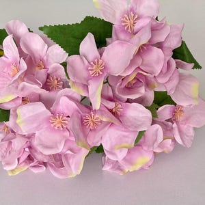 May include: A bouquet of artificial pink flowers with green leaves. The flowers have a delicate, layered appearance and are perfect for adding a touch of elegance to any space.