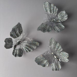 May include: Three decorative butterfly ornaments. Each butterfly has clear plastic wings dusted with silver glitter and a clear plastic body. A clear gem is set in the centre of each butterfly. The butterflies are arranged on a light grey background.