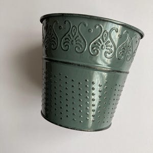 May include: A green metal planter with a decorative embossed pattern around the top rim and a dotted texture on the sides.
