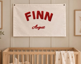 May include: A rectangular off-white fabric wall banner featuring the name FINN in large, bold, textured red letters, with the word August written below in a smaller red script font. The banner is hung by two dark metal grommets.