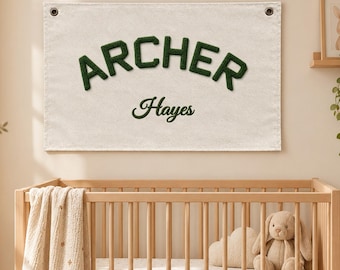 May include: A rectangular off-white fabric wall banner featuring the name Archer in dark green textured block letters and the name Hayes in dark green cursive script. The banner has two metal grommets at the top corners for hanging.
