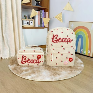 May include: A beige backpack and matching lunch bag, decorated with red strawberry motifs and the name "Becca" in red script. The backpack has a front pocket and adjustable straps. The bags sit on a small, round, fluffy rug.