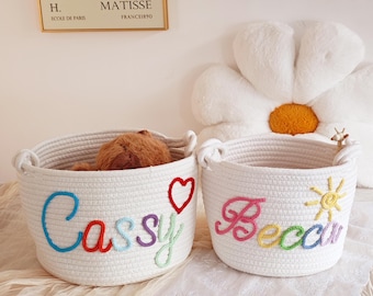May include: Two white rope storage baskets with colorful embroidered names. The basket on the left says "Cassy" with a red heart and the basket on the right says "Becca" with a yellow sun.