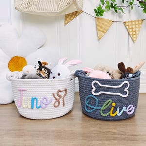 Könnte beinhalten: Zwei Seilkörbe gefüllt mit Stofftieren. Ein weißer Korb mit dem Namen "June 87" in Pastellfarben. Der andere graue Korb hat eine Knochenform und den Namen "Olive" in Pastellfarben.