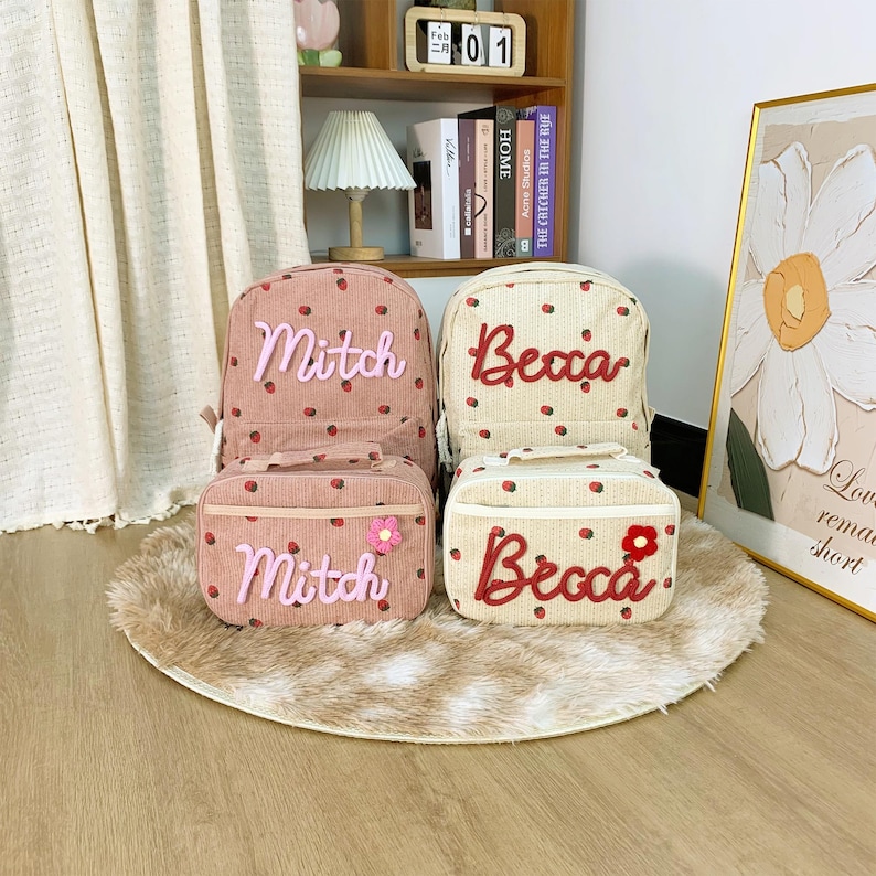 May include: A collection of personalised backpacks and lunch bags. One set is pink with a strawberry pattern and the name "Mitch". The other set is beige with a strawberry pattern and the name "Becca". Each set includes a backpack and a matching lunch bag.