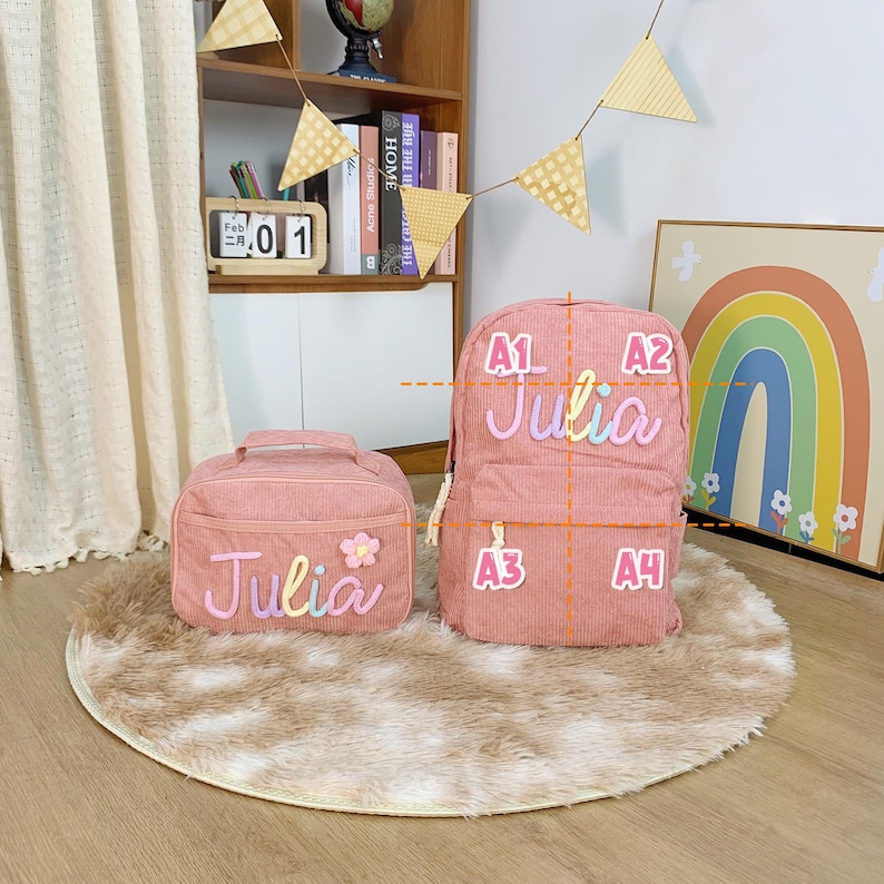 May include: A pink corduroy backpack and matching lunch bag. Both items feature the name "Julia" in colourful, embroidered lettering. The lunch bag has a flower appliqu&eacute;. The backpack has a zip closure and adjustable straps.