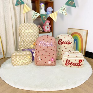 May include: A selection of personalised backpacks and lunch bags. The backpacks feature names such as "Lydia", "Mitch", and "Becca" in cursive script. Designs include a tan and white checkerboard pattern, pink corduroy with red dots, and a beige dotted pattern.