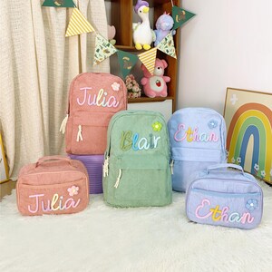 May include: A selection of pastel-coloured corduroy backpacks and lunch bags. The backpacks are pink, green, and blue, each embroidered with a name: "Julia", "Blair", and "Ethan". The lunch bags coordinate with the backpacks and feature the same names.