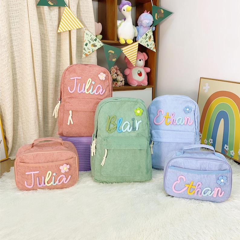 May include: A selection of pastel-coloured corduroy backpacks and lunch bags. The backpacks are pink, green, and blue, each embroidered with a name: "Julia", "Blair", and "Ethan". The lunch bags coordinate with the backpacks and feature the same names.