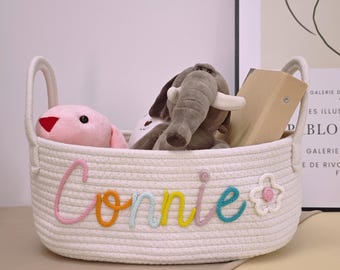 May include: White rope storage basket with the name "Connie" embroidered in colorful yarn. The basket is filled with stuffed animals, including a pink bunny and a gray elephant.