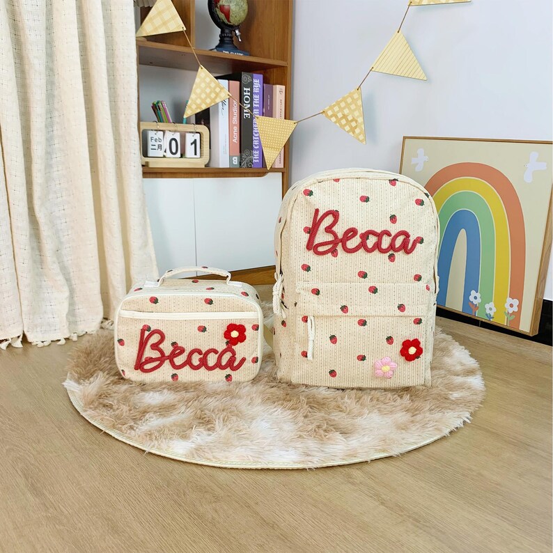 May include: A beige backpack and matching lunch bag, decorated with red strawberry motifs and the name "Becca" in red script. The backpack has a front pocket and adjustable straps. The bags sit on a small, round, fluffy rug.