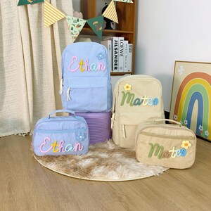 May include: A selection of corduroy backpacks and lunch bags in light blue and beige. The bags are personalised with names in colourful embroidery and decorated with flower accents. The bags are arranged on a rug.