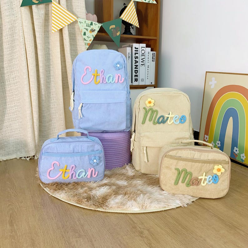 May include: A selection of corduroy backpacks and lunch bags in light blue and beige. The bags are personalised with names in colourful embroidery and decorated with flower accents. The bags are arranged on a rug.