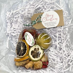 Può includere: Un sacchetto di cellophane trasparente pieno di pot-pourri di frutta secca, tra cui fette d'arancia e agrumi verdi. Un'etichetta marrone con "Merry Christmas" e "Handmade Pot Pourri" è attaccata con spago verde e bianco. Un piccolo adesivo recita "Seasons Greetings."